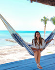 Woman sitting in a hammock on a beach with palm trees and blue water in the background.