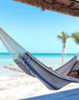 Person relaxing in a blue and white patterned hammock on a tropical beach.