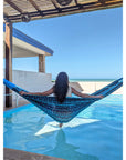 Person relaxing in a blue hammock by a pool with a clear blue sky.