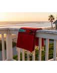 Red blanket with fringe draped over a railing overlooking a beach at sunset.
