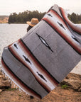 Person sitting by a lake with a gray blanket featuring black Mexican designs and stripes.