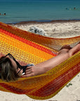 Person relaxing in a colorful hammock on a sandy beach with people in the water.