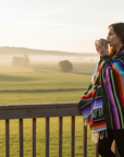 woman on a deck wearing a blanket sipping from a mug, looking out over farmland.