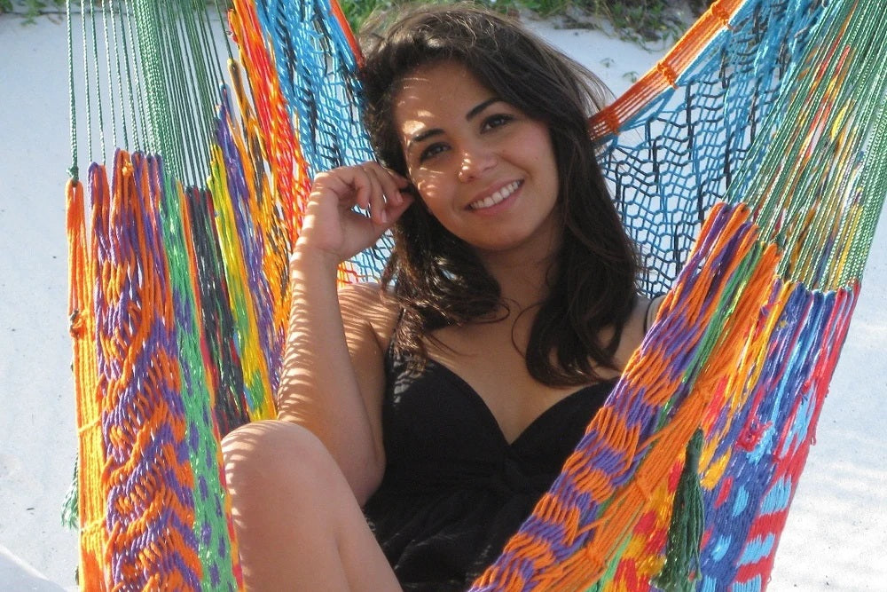 A woman smiling while sitting in a brightly colored woven hammock on a white sandy beach.