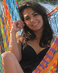 A woman smiling while sitting in a brightly colored woven hammock on a white sandy beach.
