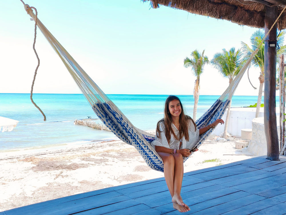 Woman sitting in a hammock on a beach with palm trees and blue water in the background.