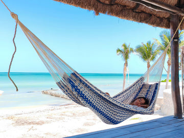 Person relaxing in a blue and white patterned hammock on a tropical beach.