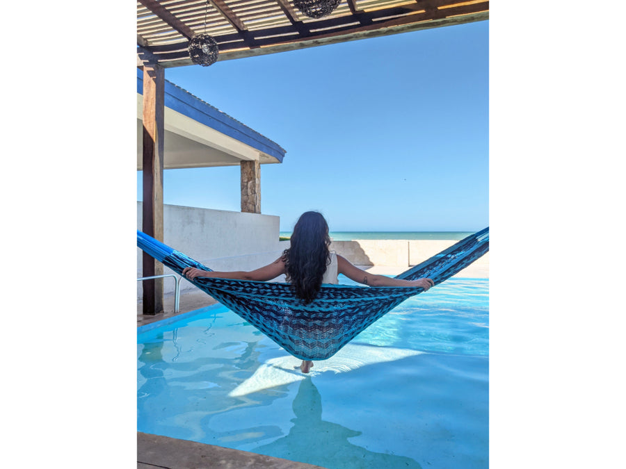 Person relaxing in a blue hammock by a pool with a clear blue sky.