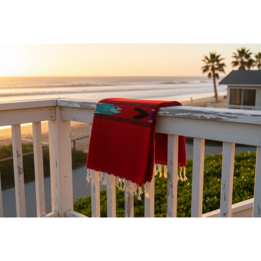 Red blanket with fringe draped over a railing overlooking a beach at sunset.