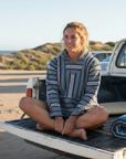Woman sitting on a truck bed by the beach wearing a grey hoodie.