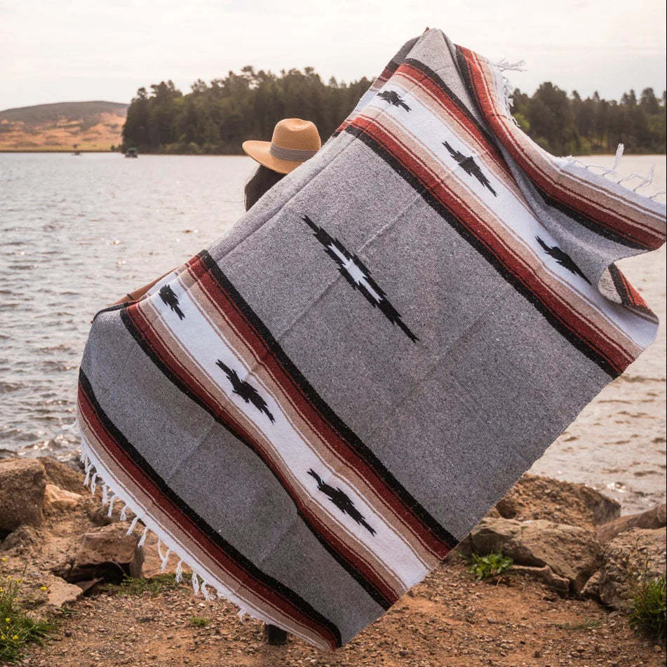 Person sitting by a lake with a gray blanket featuring black Mexican designs and stripes.