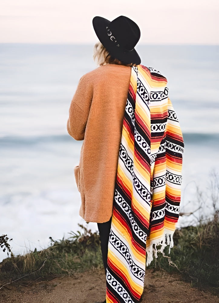 Woman looking out over the ocean with a blanket draped over her shoulderand wearing a black hat
