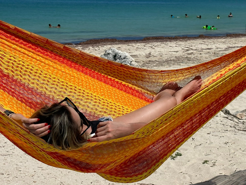 Person relaxing in a colorful hammock on a sandy beach with people in the water.