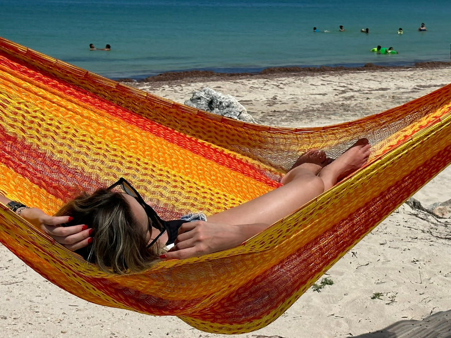 Person relaxing in a colorful hammock on a sandy beach with people in the water.
